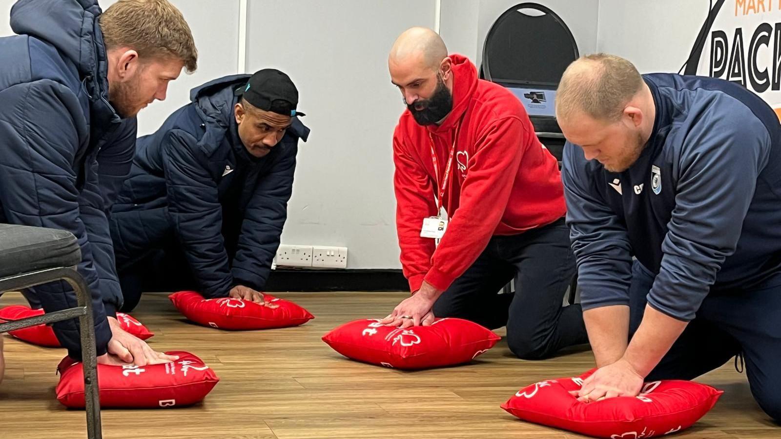 Four men are in a room, kneeling down, each simulating chest compressions on a red cushion. Three of them are in Cardiff Rugby training kit. The fourth man is wearing a red British Heart Foundation jumper. The floor is wooden and in the background there are white walls with stacked chairs and plug sockets.