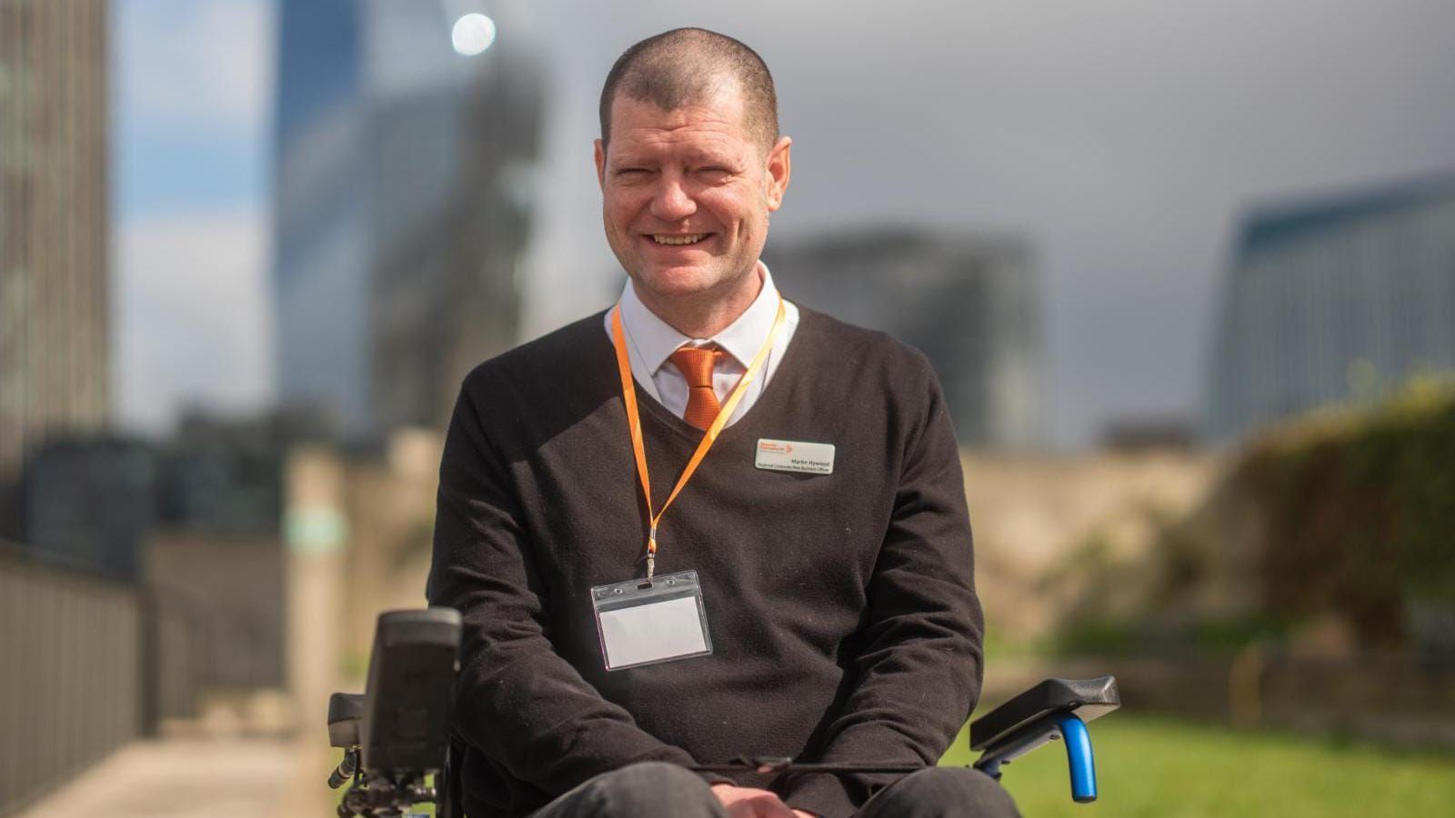 Martin Hywood sitting in a wheelchair while wearing a jumper, white shirt and red tie. He is smiling at the camera 