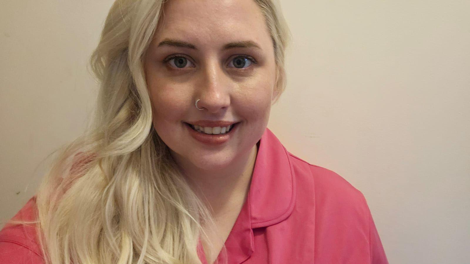 Sophie has wavy blond hair which is pulled over her right shoulder. She is staring directly into the camera wearing a pink work uniform.