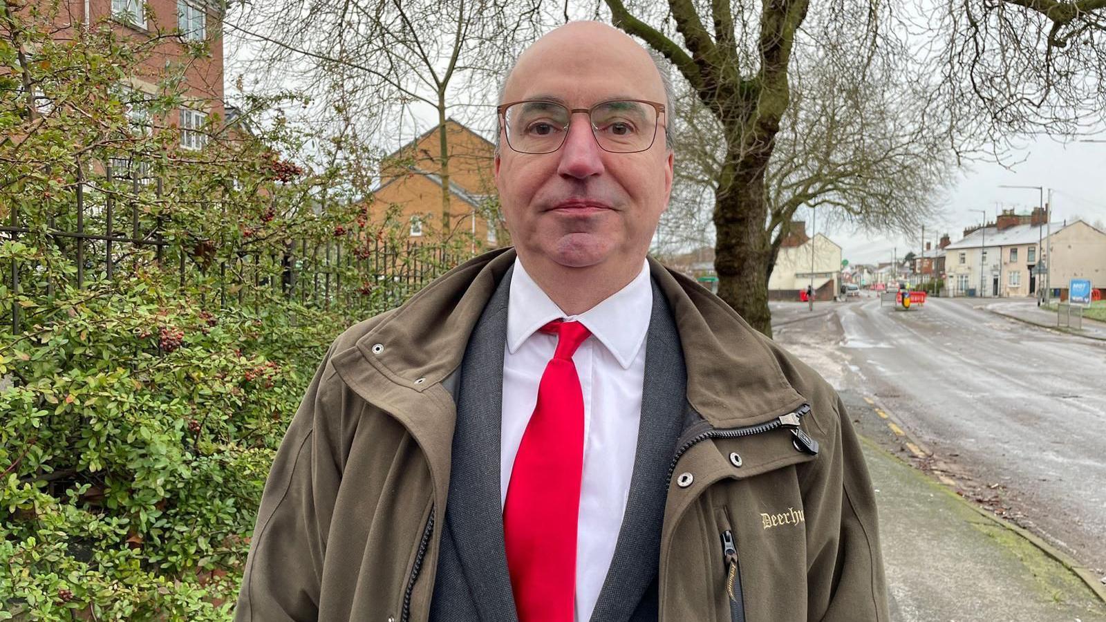 A middle-aged bald man is wearing a white shirt, a bright red tie and a grey jacket underneath a brown coat. He is standing on a quiet street with trees and bushes in the background.