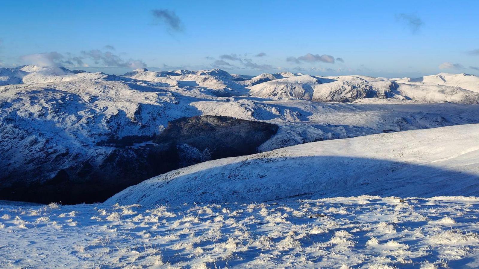 The view from the summit of a snowy Helvellyn looking over the southern and western fells. Snow carpets the peaks in the skyline as the sunrise breaks over the horizon.