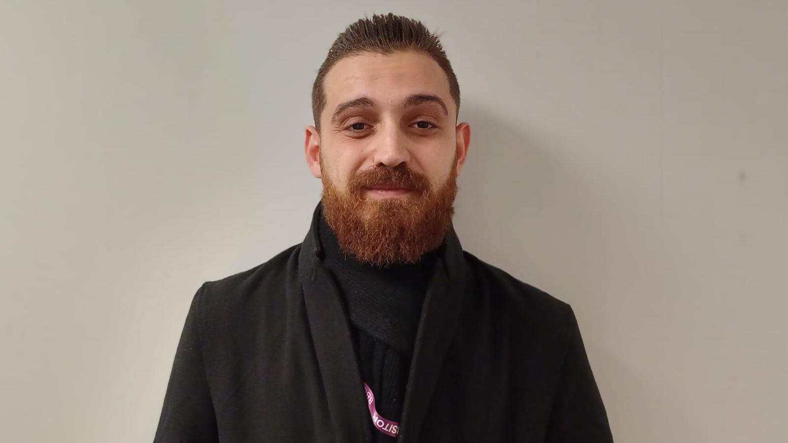 Mohammed is standing against a white background. He's wearing a black coat and scarf, and looking into the camera. He's a beard.