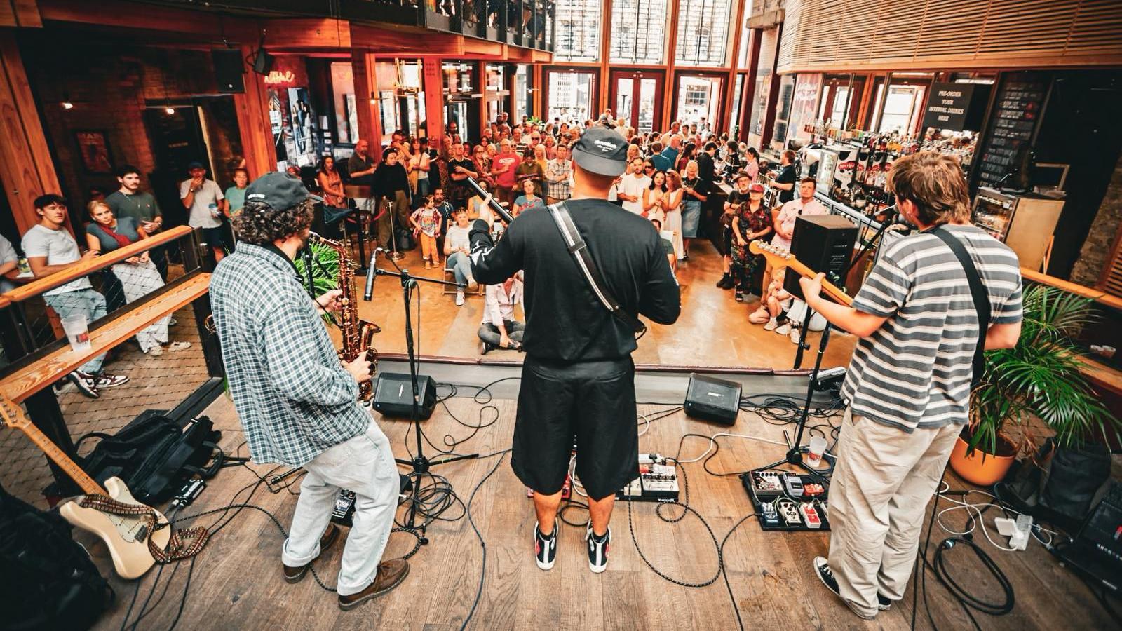 Three male musicians, their backs to the crowd, play at Old City Sounds in Bristol. They are standing on a wooden stage and beyond them is a crowd of dozens of people watching on