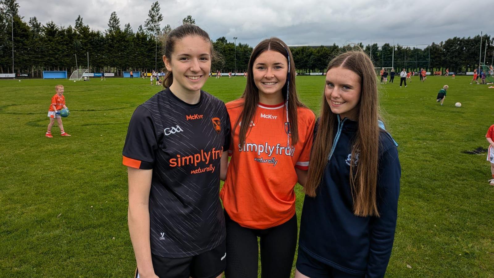 Three girls standing with their arms round each other on a GAA pitch