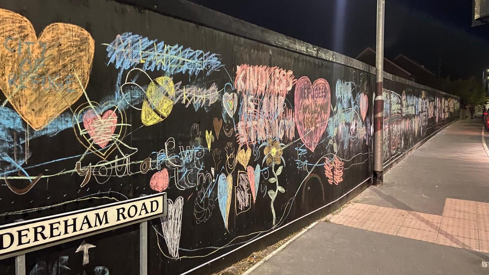 A picture which has been taken at night of the hoarding which has been covered in chalk messages of support. However, the messages have all been crossed out. There is also a road sign on the pavement which says, DEREHAM ROAD.