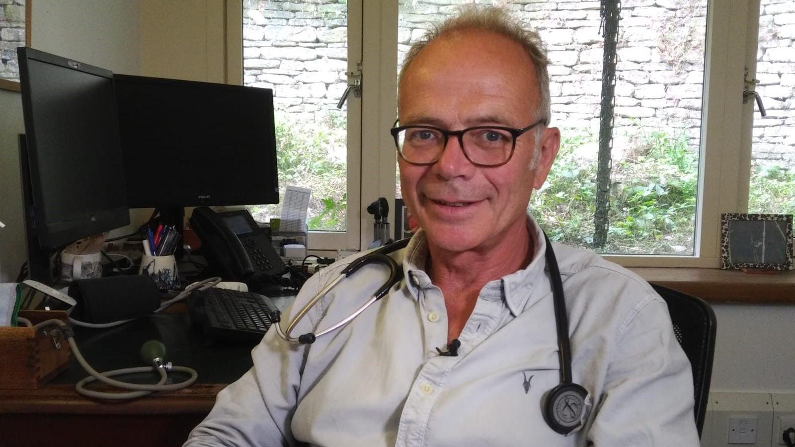 Dr Simon Opher with a stethoscope around his neck, sitting in front of a desk