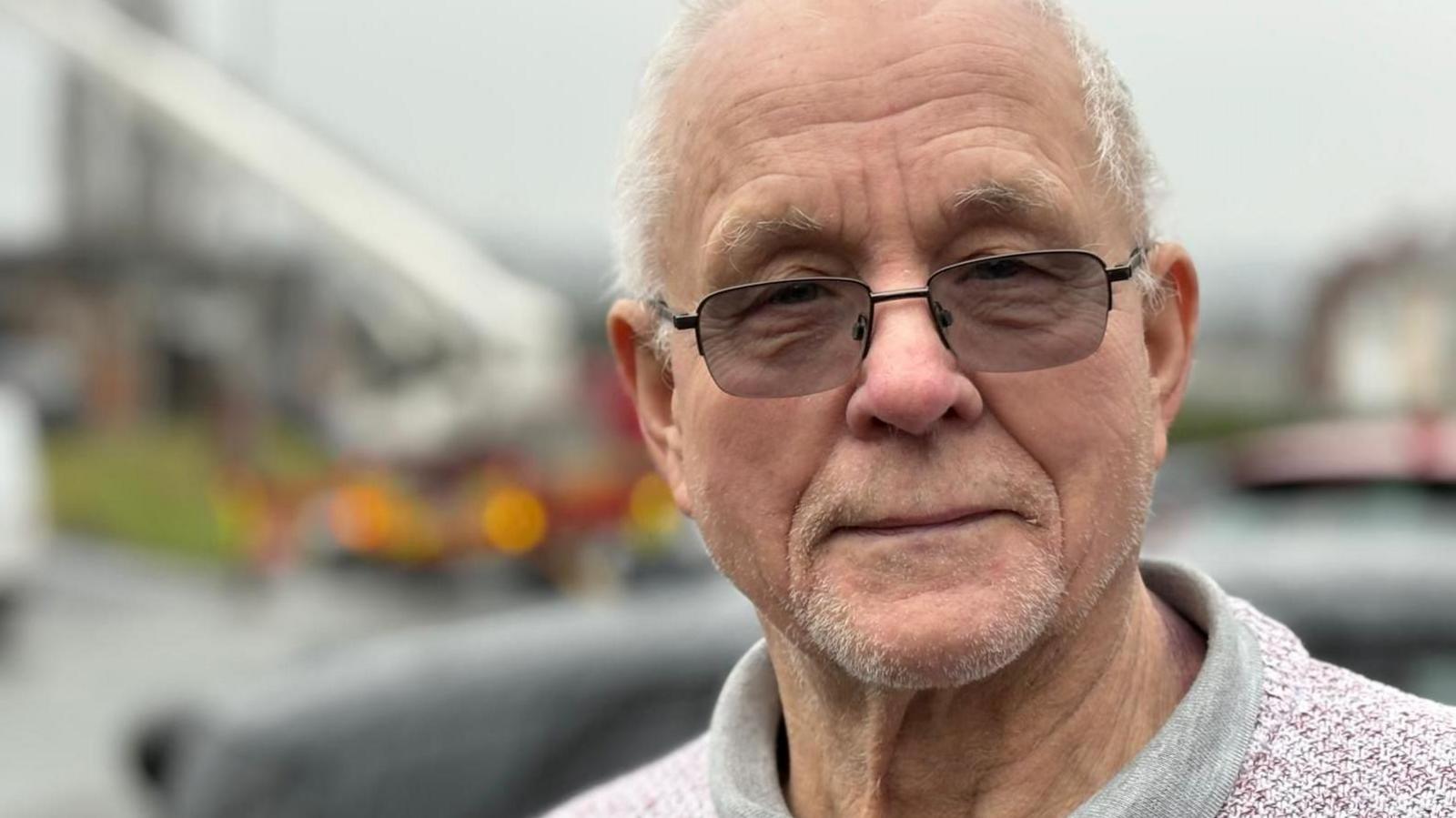 A head shot of a man who is wearing a jumper and looking at the camera. He is wearing glasses and has grey hair. 