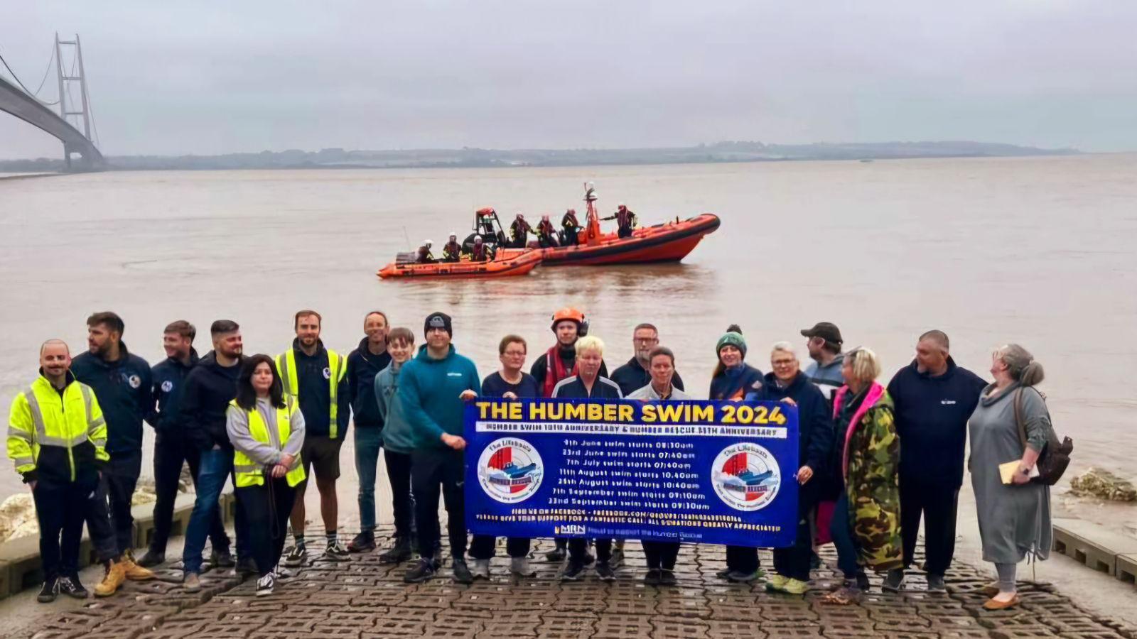 Twenty men and women stood in front of the River Humber on pavement holding a blue and yellow sign reading 'The Humber Swim 2024'. Most of them are dressed in blue uniform and some of them have hi-vis vests on. In the background are two Humber Rescue orange lifeboats with volunteers on board. 