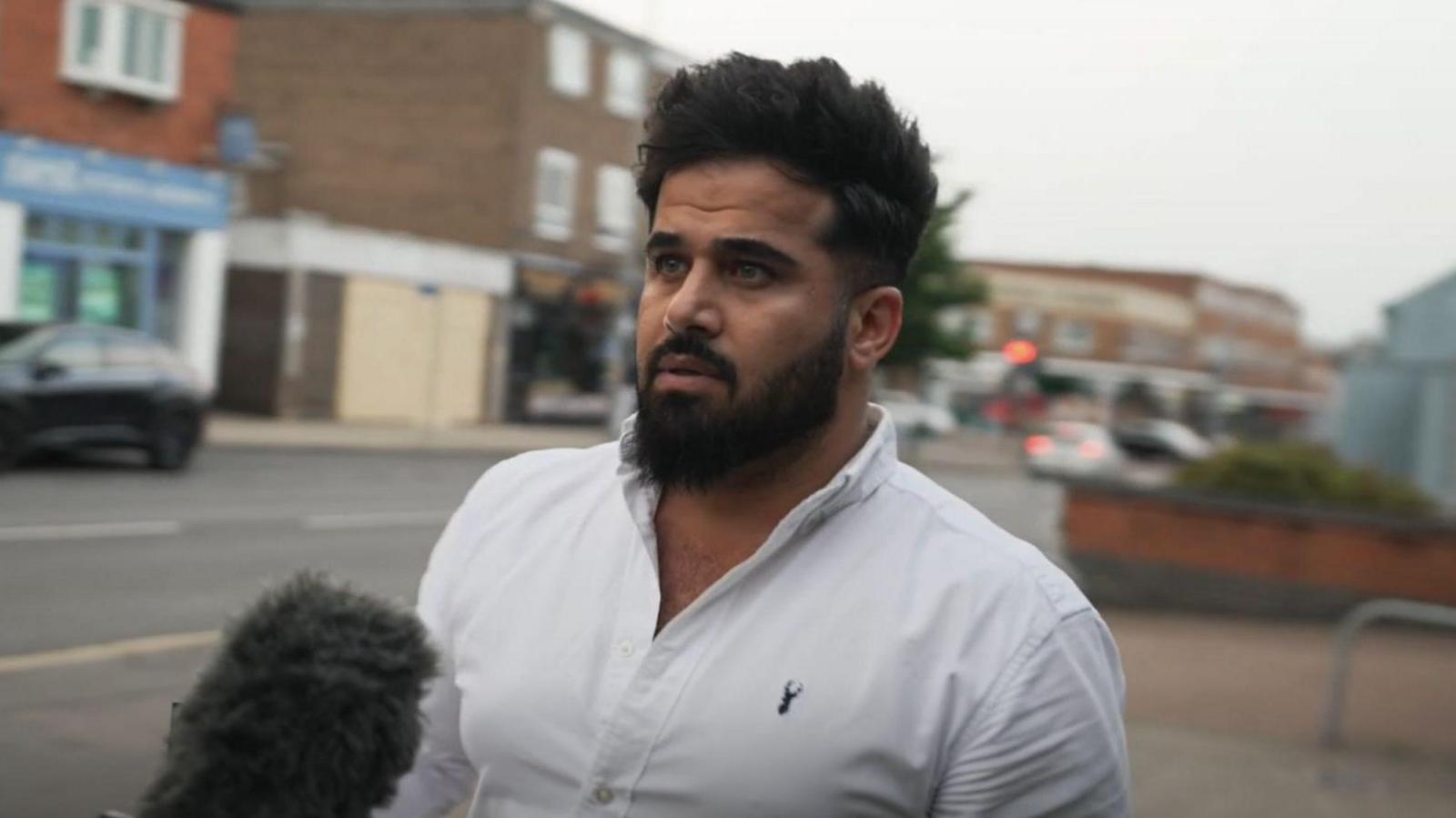 A picture of a man with dark hair and a beard. He wears a white shirt.