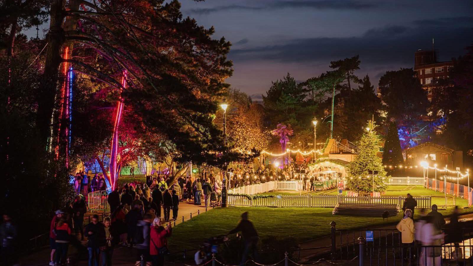 Bournemouth's Christmas train ride in Lower Gardens at night. The park trees are illuminated. The path through the middle is full of people. The mini train ride is fenced of with white picket fencing. The track circles a large tree and goes through a lit up tunnel station.