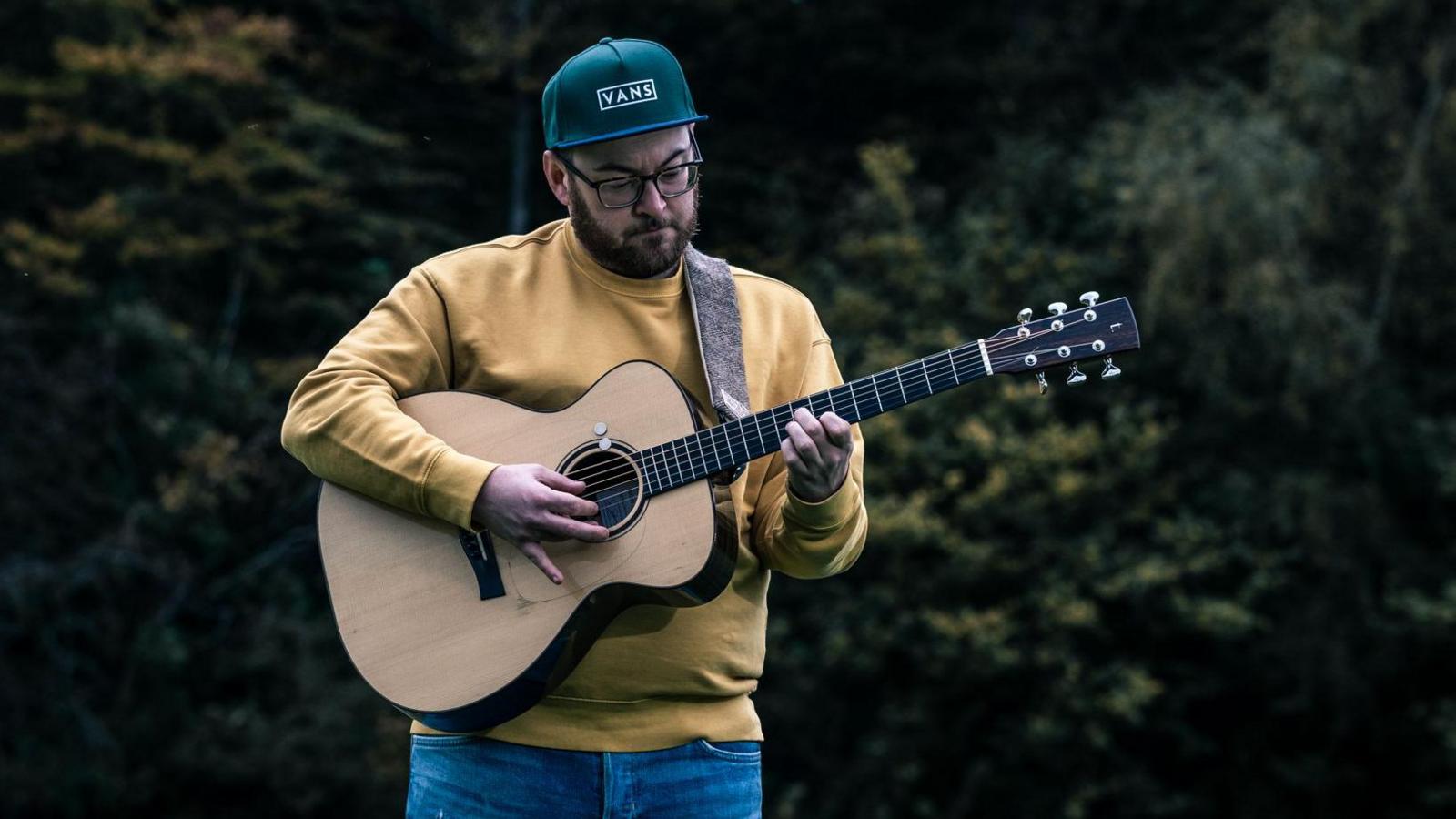 A man holding an acoustic guitar with some trees in the background. He is wearing a beige jumper and a green Vans baseball cap. He is wearing glasses and is dark-haired with a beard. 