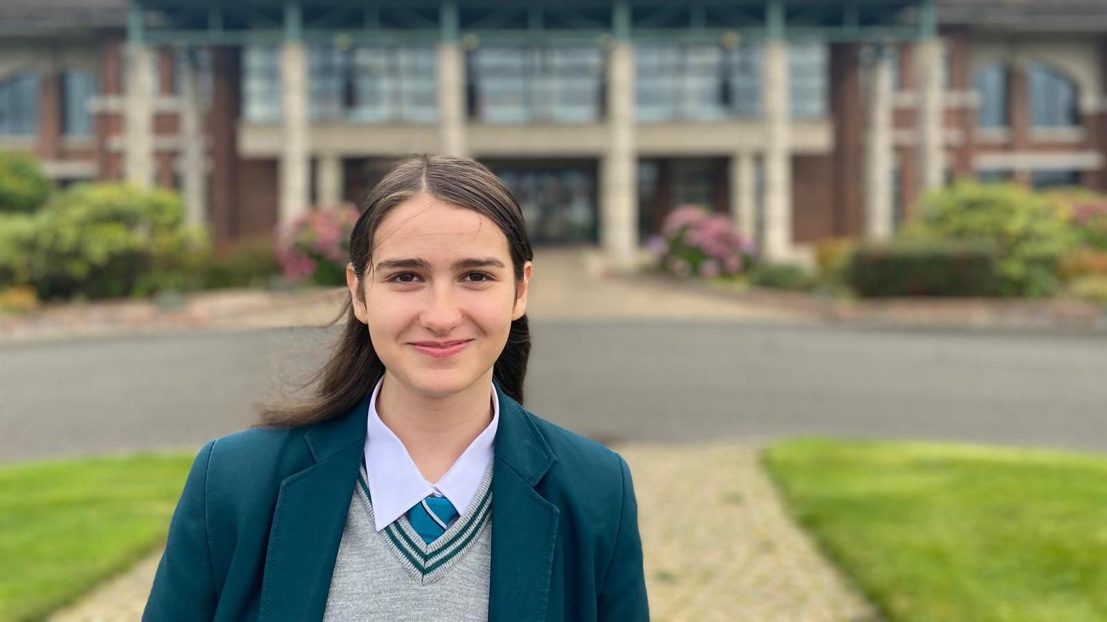 Estela wears her school uniform -a teal jacket with grey jumper, teal stripe round the collar and teal tie. She has brown her half up half down. 