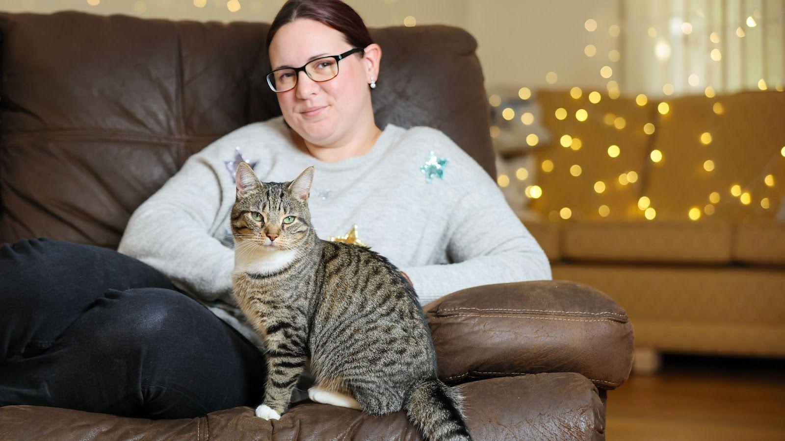 A woman is curled up on the sofa, smiling at the camera, with a tabby cat in front of her, there are Christmas lights in the background