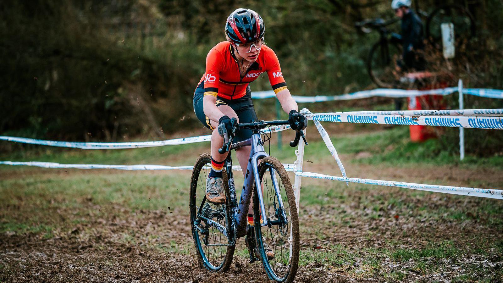 Lauren is seen riding her bike, which is covered in mud. She wears cycling shorts, an orange top and helmet as well as goggles. 