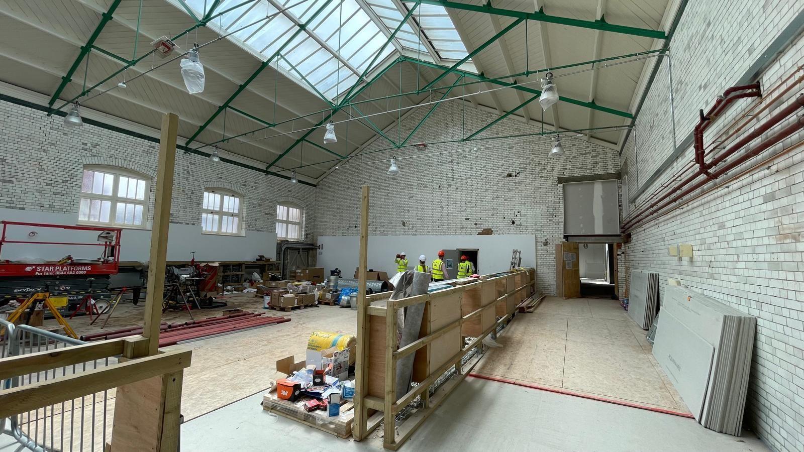 A large, tall room which looks very industrial. There are white glazed bricks covering every wall and big cream rectangular windows. The room is full of tools and wooden boards. The room is in the process of being sectioned off. To the left of the partition will be the gym.