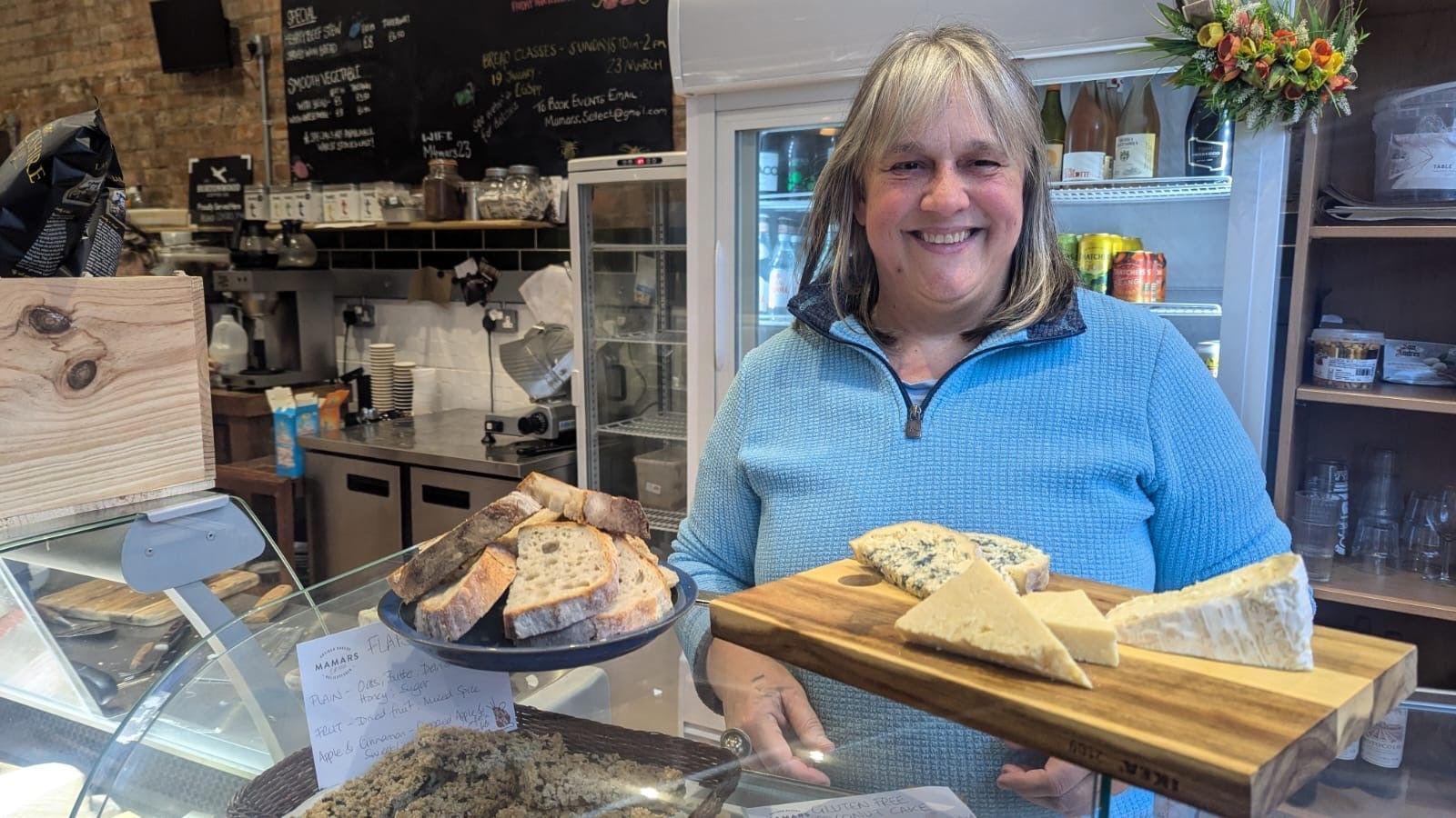 Sonia Johnson smiling wearing a blue top, while stood in her bakery behind the bread counter