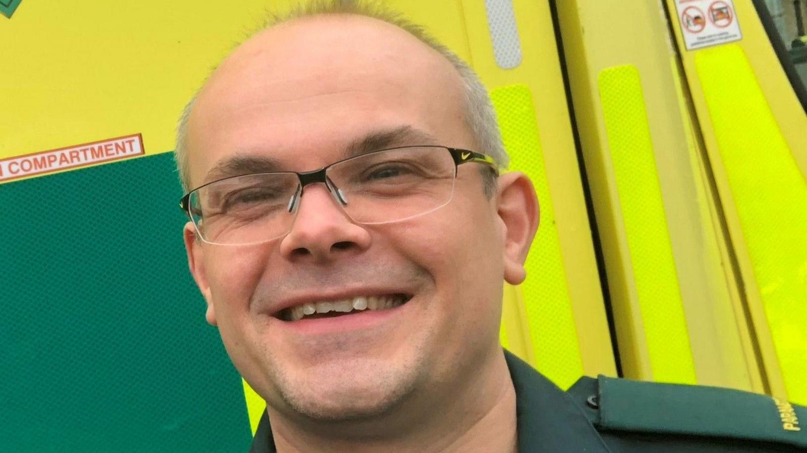 Professor John Martin, with short hair, wearing glasses, smiling, looking straight at the camera, wearing a paramedic green uniform and standing in front of an yellow ambulance