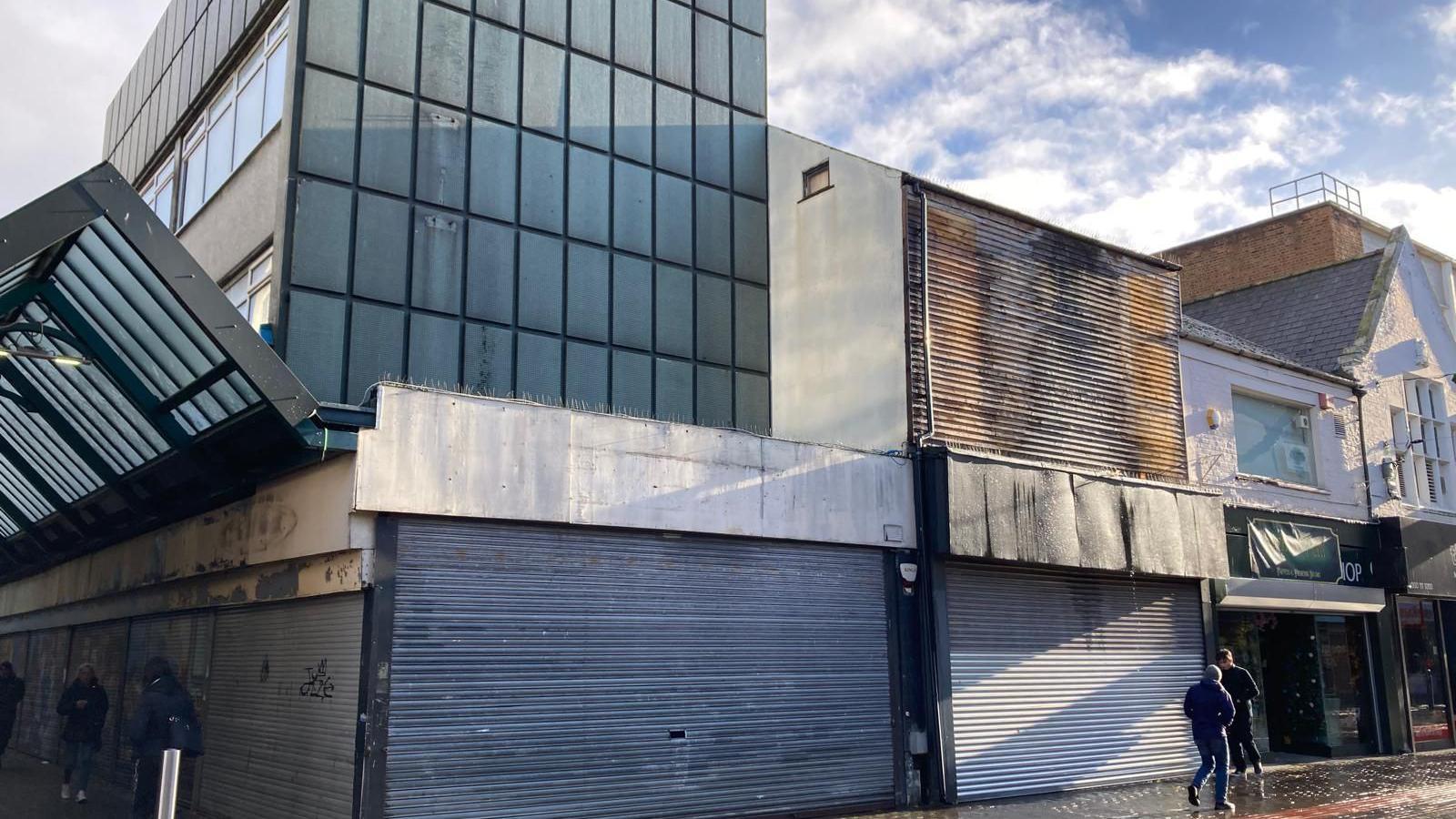 The blue shutter of the former H. Samuel in Middlesbrough are down and the upper floors of the grey-panel building are also empty. The store next door is closed too. Two people are walking past.