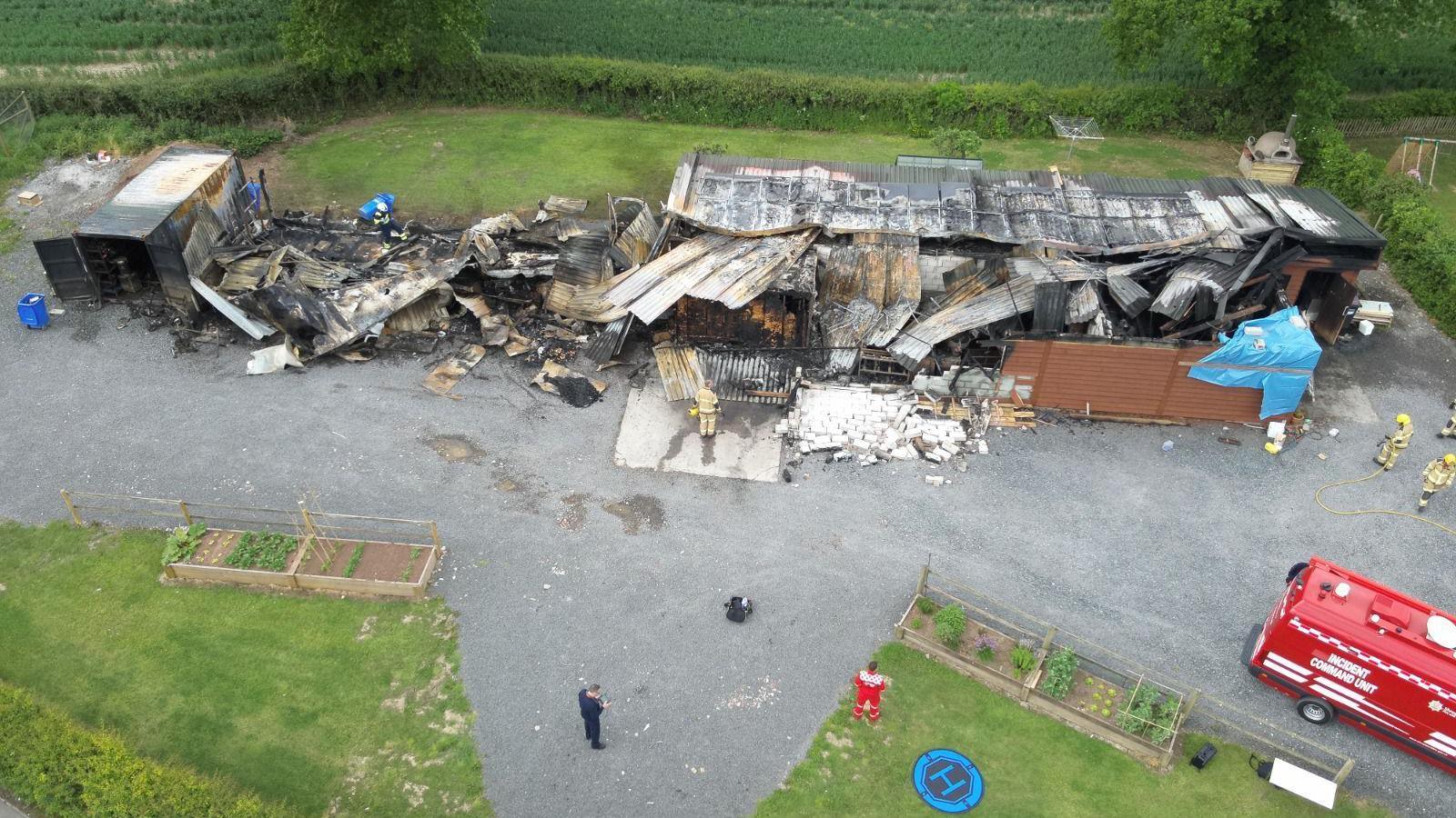 An aerial photograph shows a destroyed building with a charred and collapsed roof. Firefighters in beige and yellow uniforms can seen around the edged of the building. A red truck can be seen on the right.