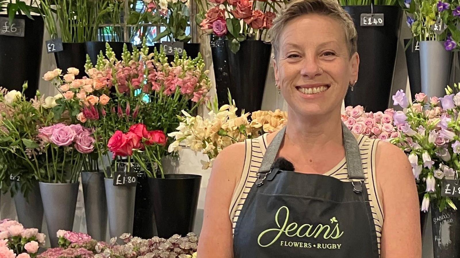 Miki Croner stands with rows of buckets containing different types of flowers behind her. The ones closest to her are all pink, cream and lilac. She is wearing an apron that says Jean's Flowers, Rugby. She has short hair and is wearing a summer, sleeveless top.