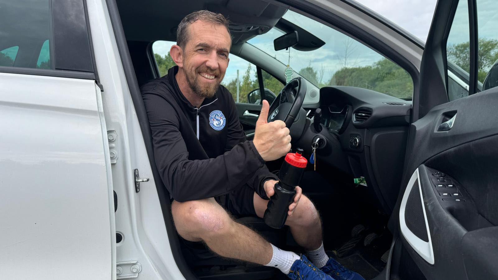 Camera Alexander sitting in the driving seat of a white vehicle with the door open. He is in sports gear and is holding a black water bottle and smiling while holding his thumb up.