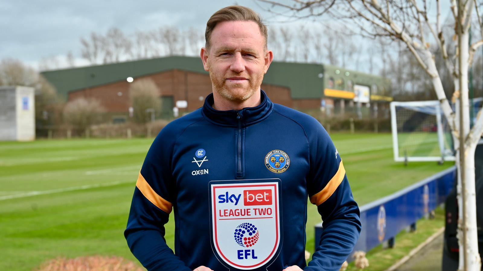 Shrewsbury Town head coach Gavin Cowan holds the League Two manager of the month award with the training ground pitch in the background