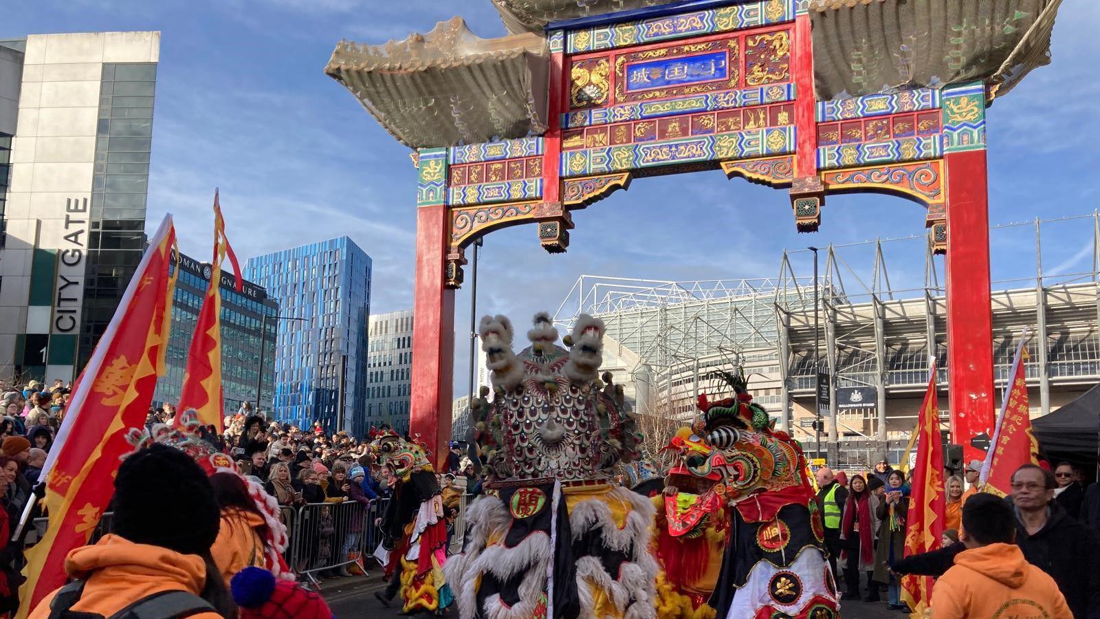 Crowds surround the ornamental gate which leads to Newcastle's Chinatown. People in elaborate costumes are walking under it .