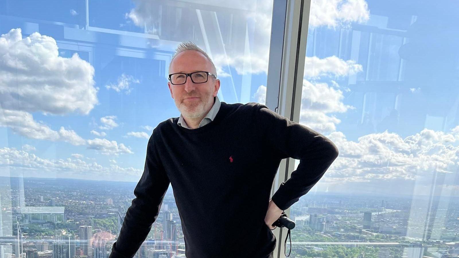 A man in a black jumper stood in front of a large glass window.
