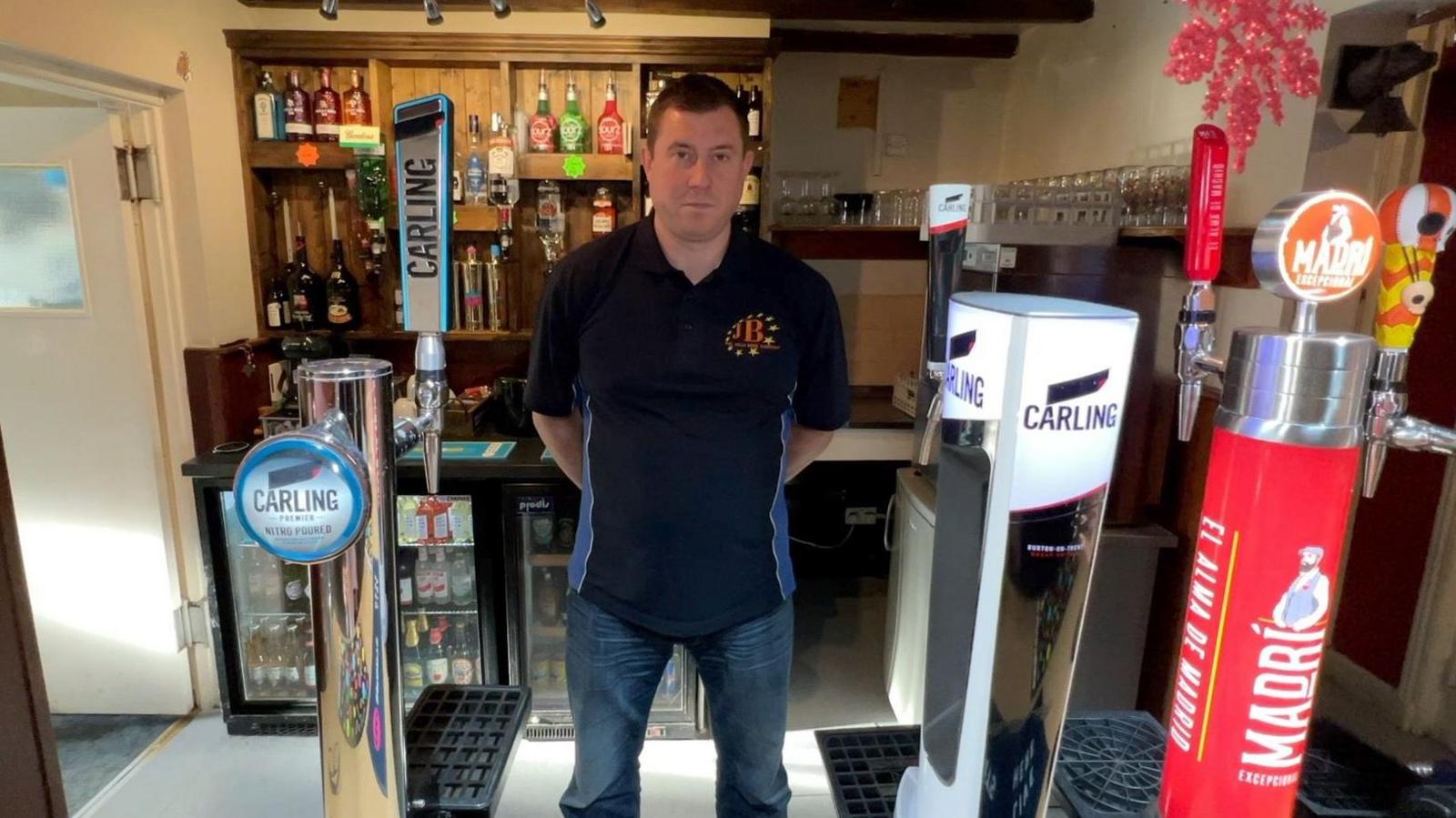 A man wearing a black-shirt stands behind a bar in a pub.