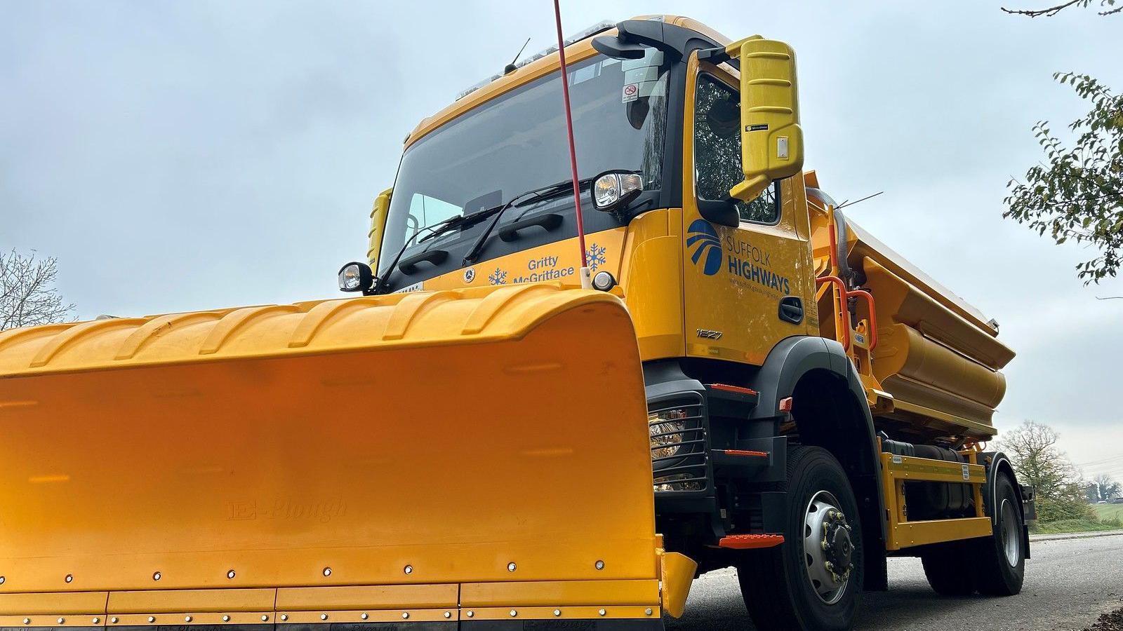 A Suffolk Highways yellow gritter vehicle with a snow plough on its front is pictured stationary on a road.