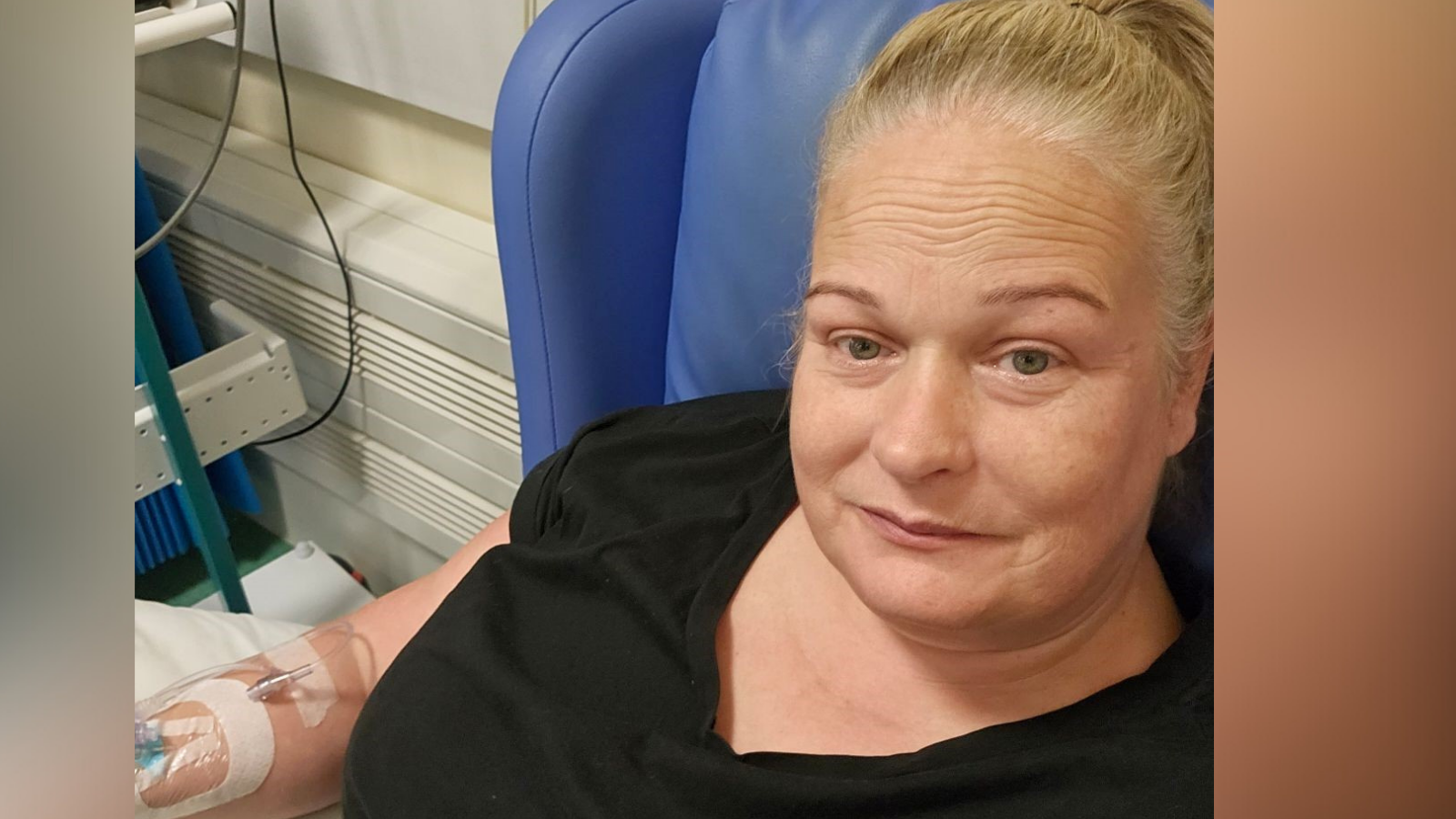 Penny has her blond hair tied up and is sitting on a blue chair. She has an IV in her right arm. 