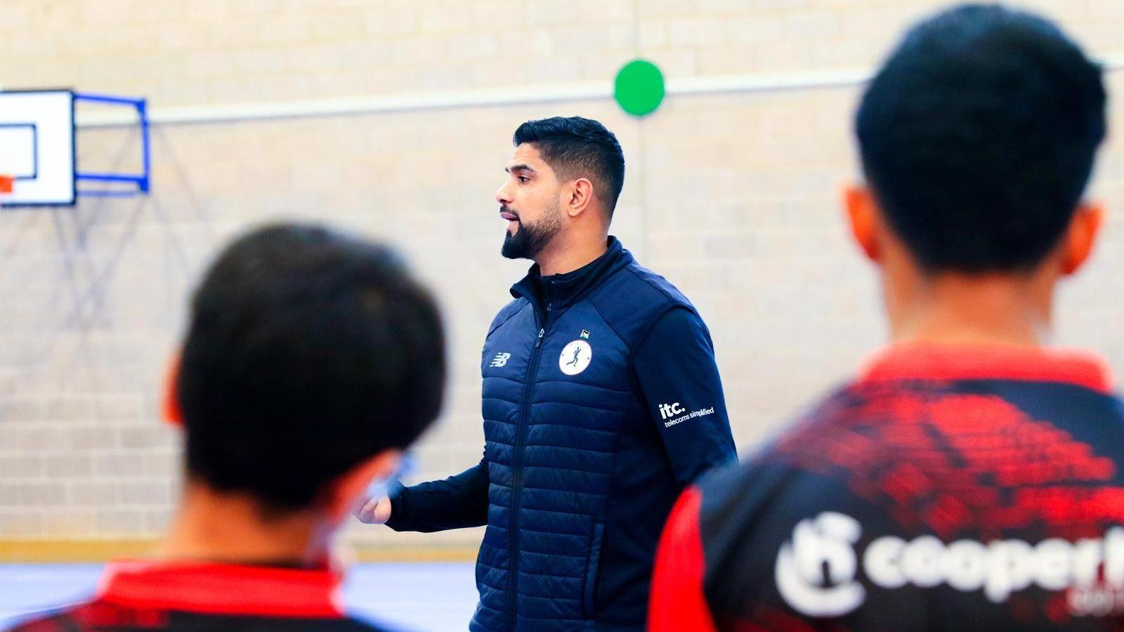 A man wearing a blue gilet talking with the back of the heads of two boys wearing red and blue shirts with a white-lettered logo on the back. 