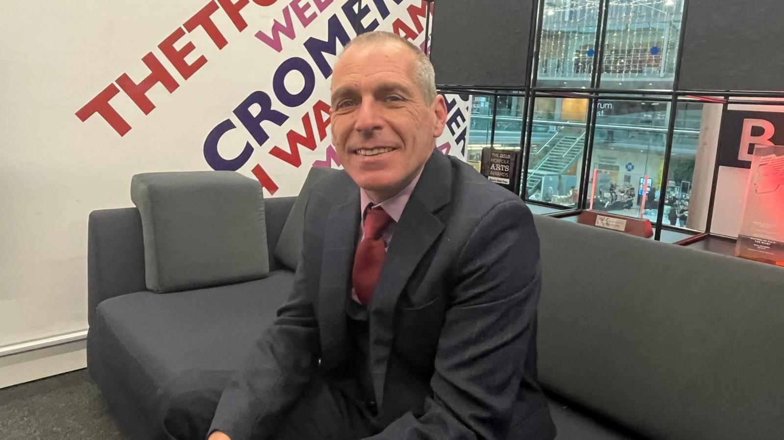 Peter Dutch is sitting on a grey sofa in a BBC studio. He is smiling at the camera and is wearing a dark suit, pink shirt and red tie.