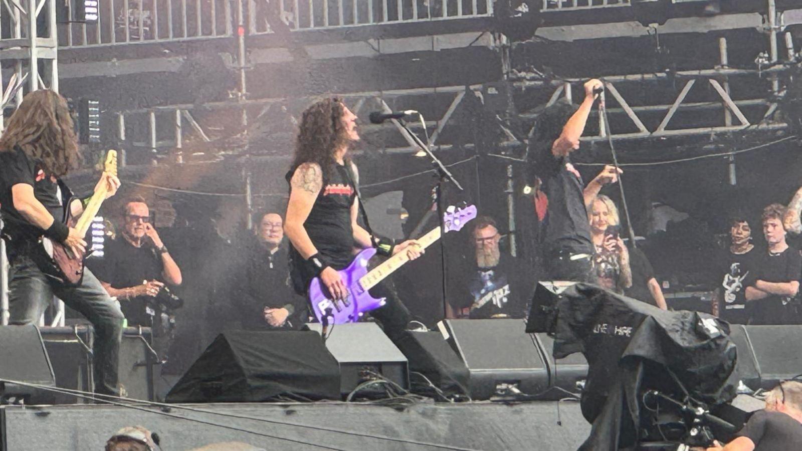 A heavy metal band perform on stage. The lead singer holds a purple guitar and sings into a microphone