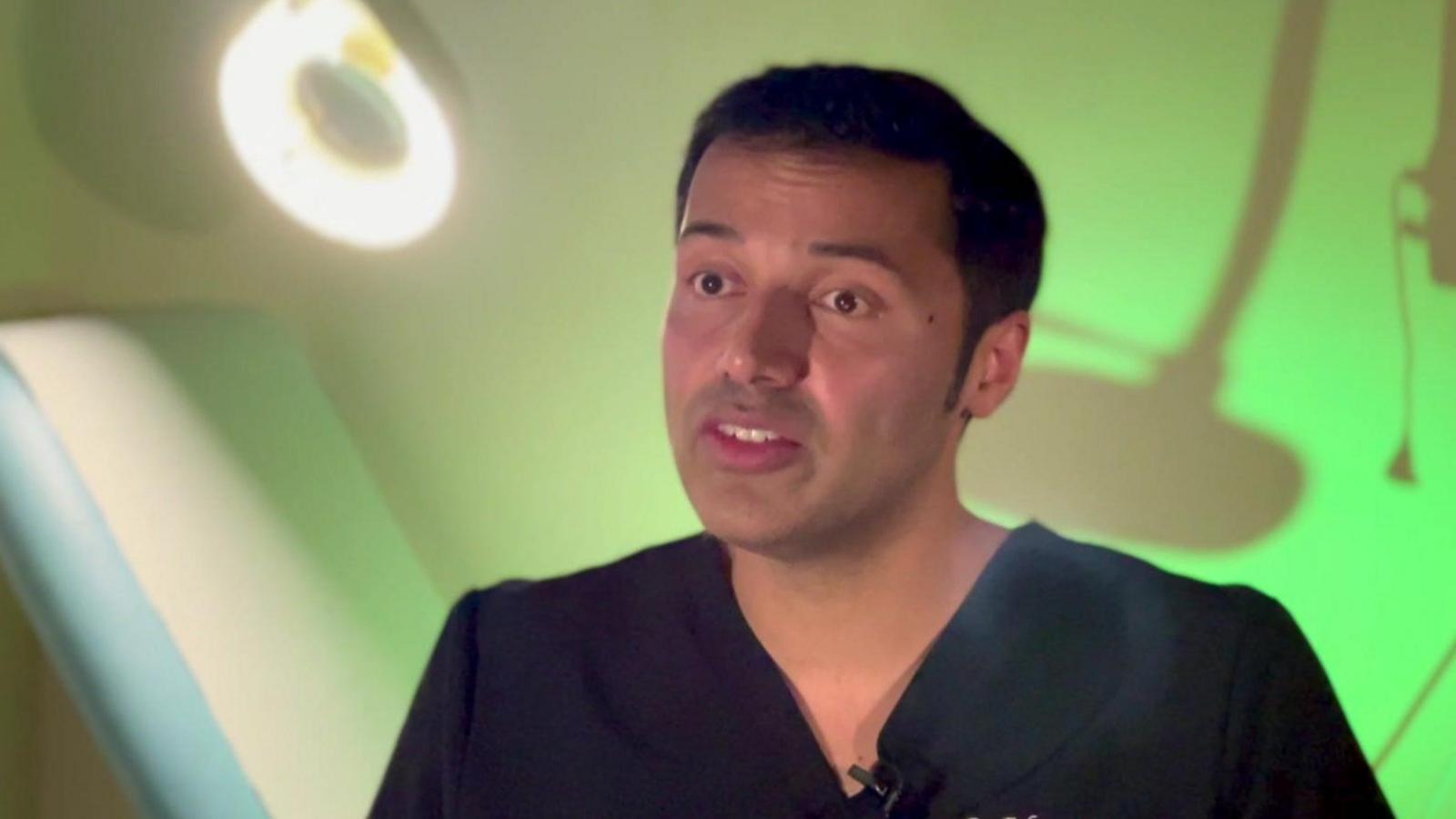 A man wearing dark blue scrubs with a medical bench in white and blue behind him.