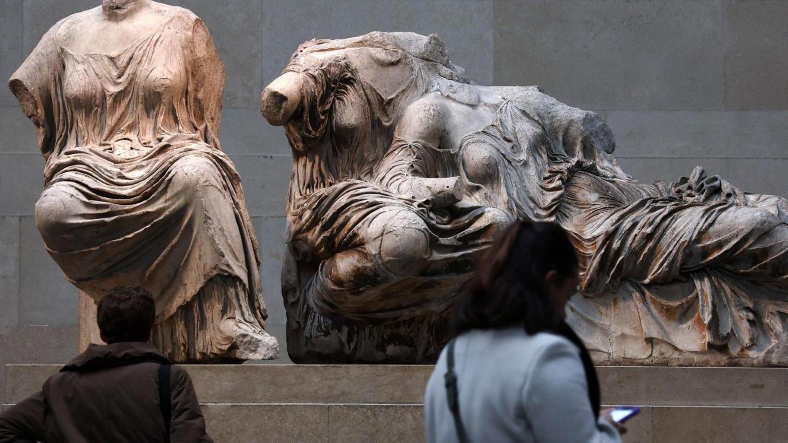 Part of the Elgin marbles are displaced with two women looking at them. One wears a brown cut and the other a pale blue coat, looking at her phone.