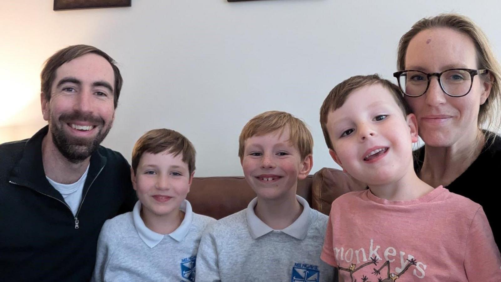 Dad Calum, his sons Archie, Andrew, Roddy and mum Katie are sat side by side on a couch smiling at the camera. Archie and Andrew are in their school uniform and Roddy is wearing a top with monkeys on it. 