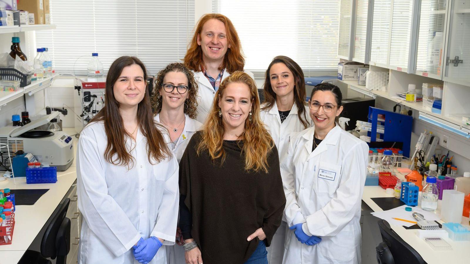 Group of six scientists - five women and a man - taking a group picture, most wearing white lab coats in a laboratory setting.