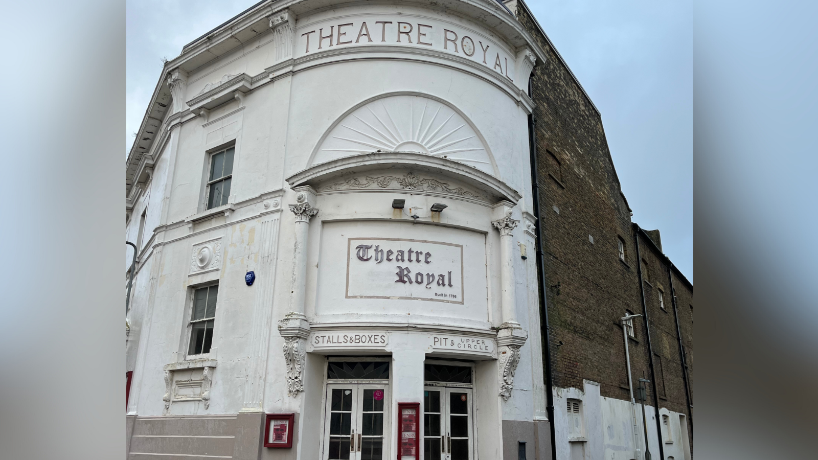 An exterior picture of the Theatre Royal in Margate. The two-storey building is white and has two doors of entry. On the left side there is a sign for 'stalls and boxes' and on the right is a sign for 'pit and upper circle'. Above those two signs is a sign stating 'Theatre Royal'. 