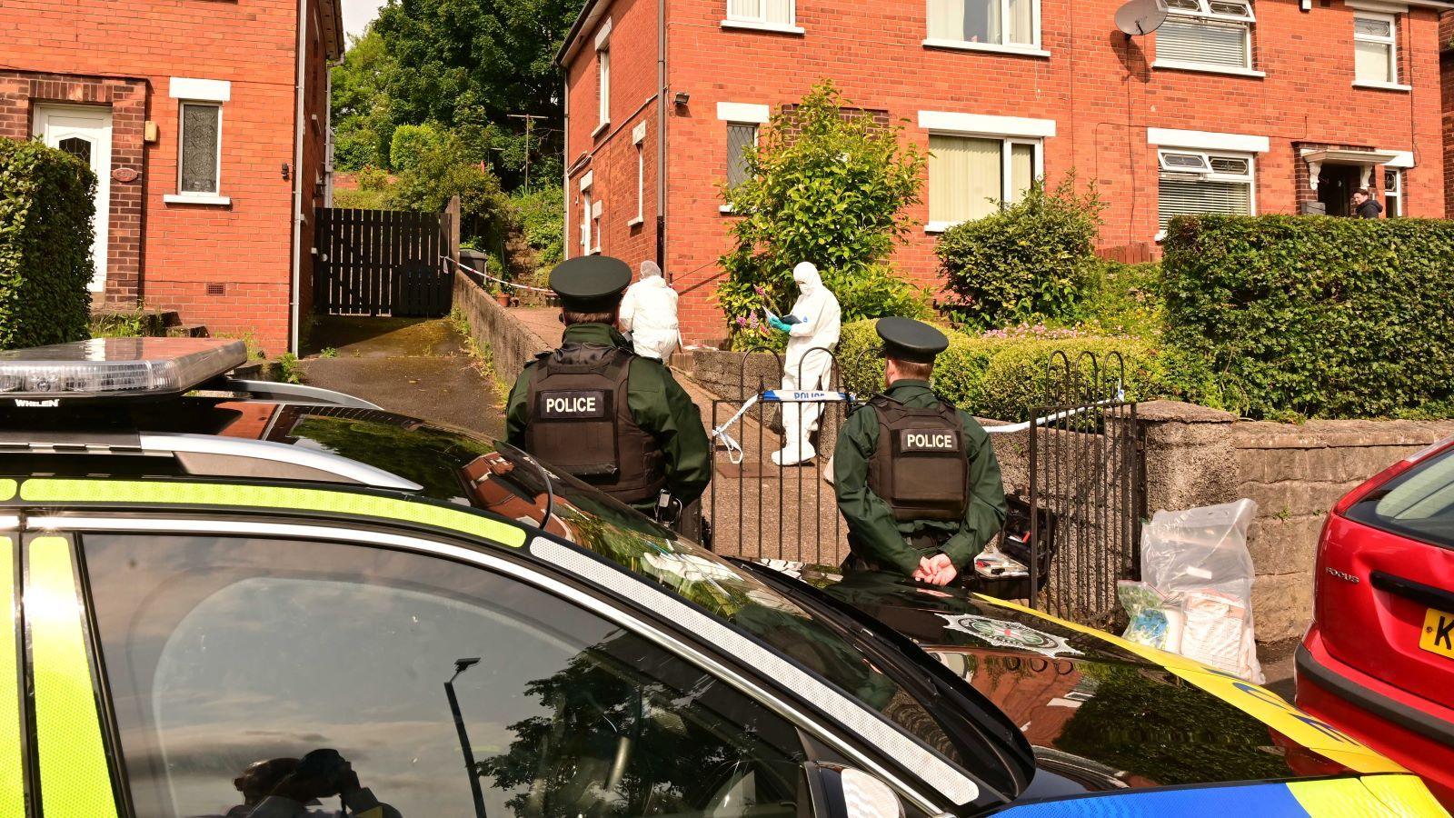 A photo taken from the opposite side of a road showing a parked police car outside a row of semi-detached, red brick houses in north Belfast. Three uniformed officers are standing with their backs to the camera. A red car is also parked blocking the driveway of the house. A cordon is placed along the bottom and mid-way up the driveway of the house.