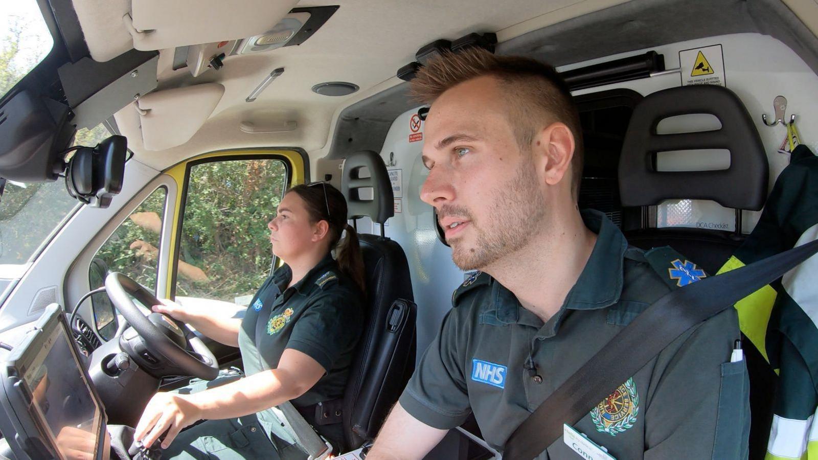 Two paramedics in their ambulance, dressed in dark green polo shirts with the NHS and ambulance service logos