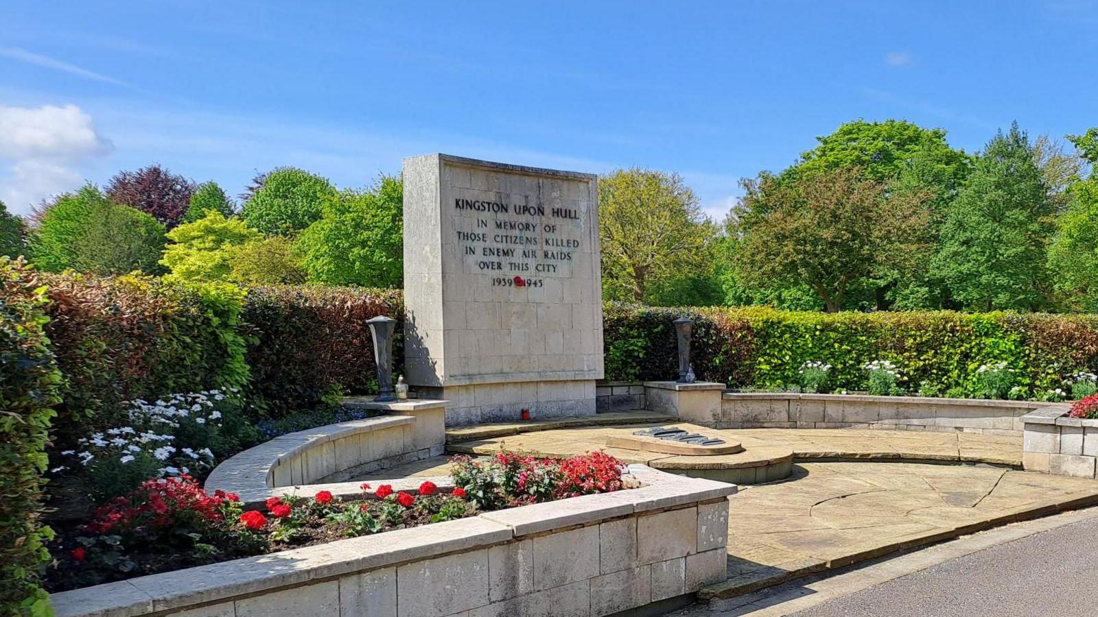 A stone memorial area. The memorial is surrounded by leafy green trees and hedges with red and white flowers planted in front of a walled area. An inscription on the memorial reads "In memory of those citizens killed in enemy air raids over this city".