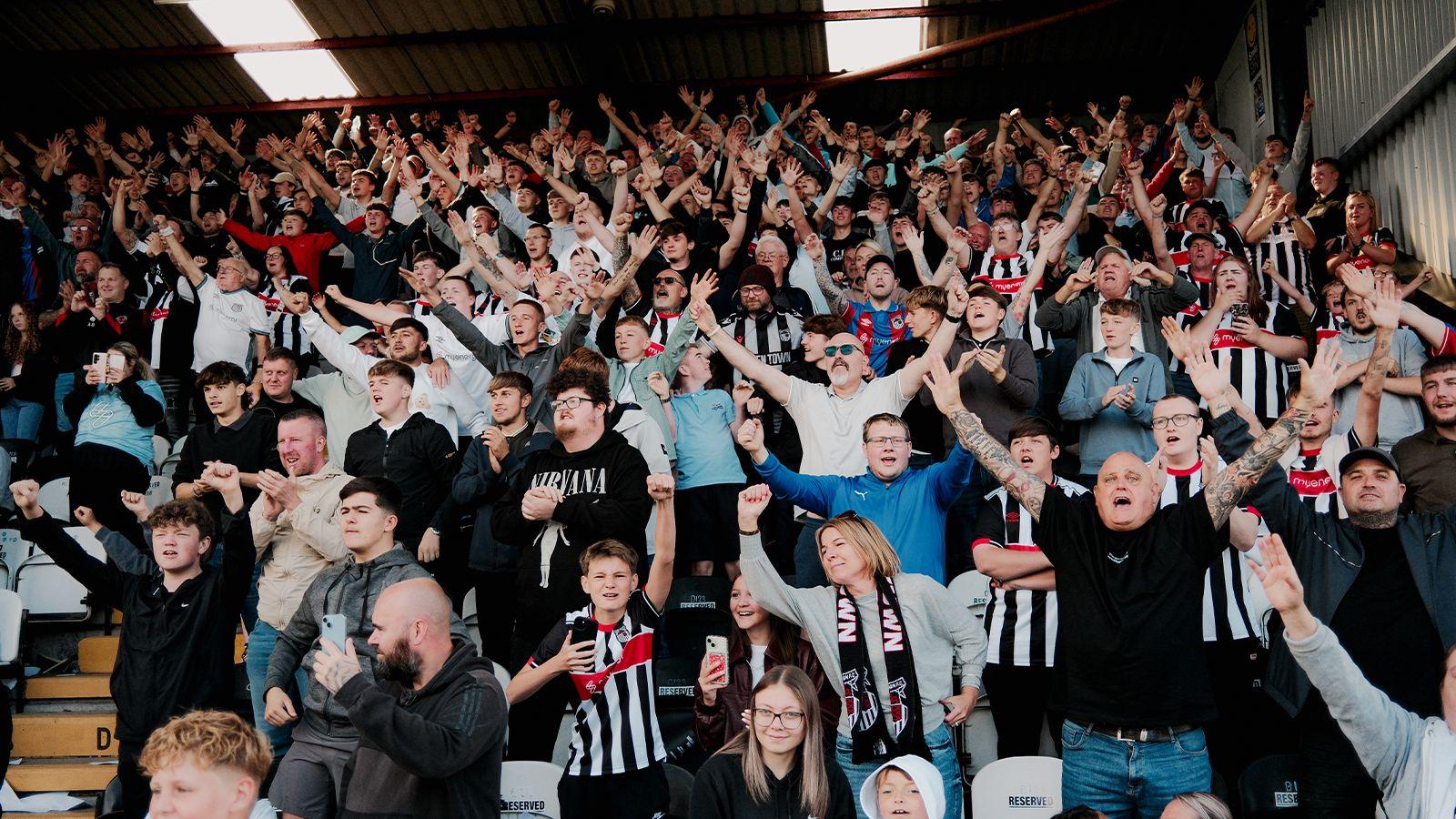 Crowd of cheering Grimsby Town fans with arms aloft, Many are wearing the club's black and white striped kit.