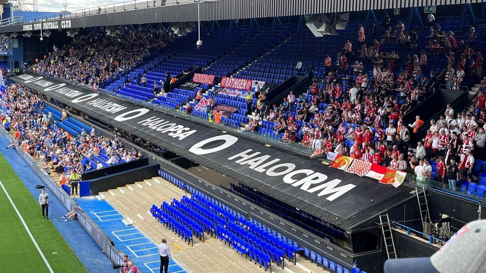 The Cobbold Stand at Portman Road. The seats are blue and there is a long black roof separating the upper and lower tiers. To the right are Fortuna Dusseldorf supporters who have filled a quarter of the upper tier. They are mostly wearing red. To the right are the Ipswich fans, who have filled the half of the stand they are allocated. A corner of the pitch can be seen in the bottom left corner of the picture.