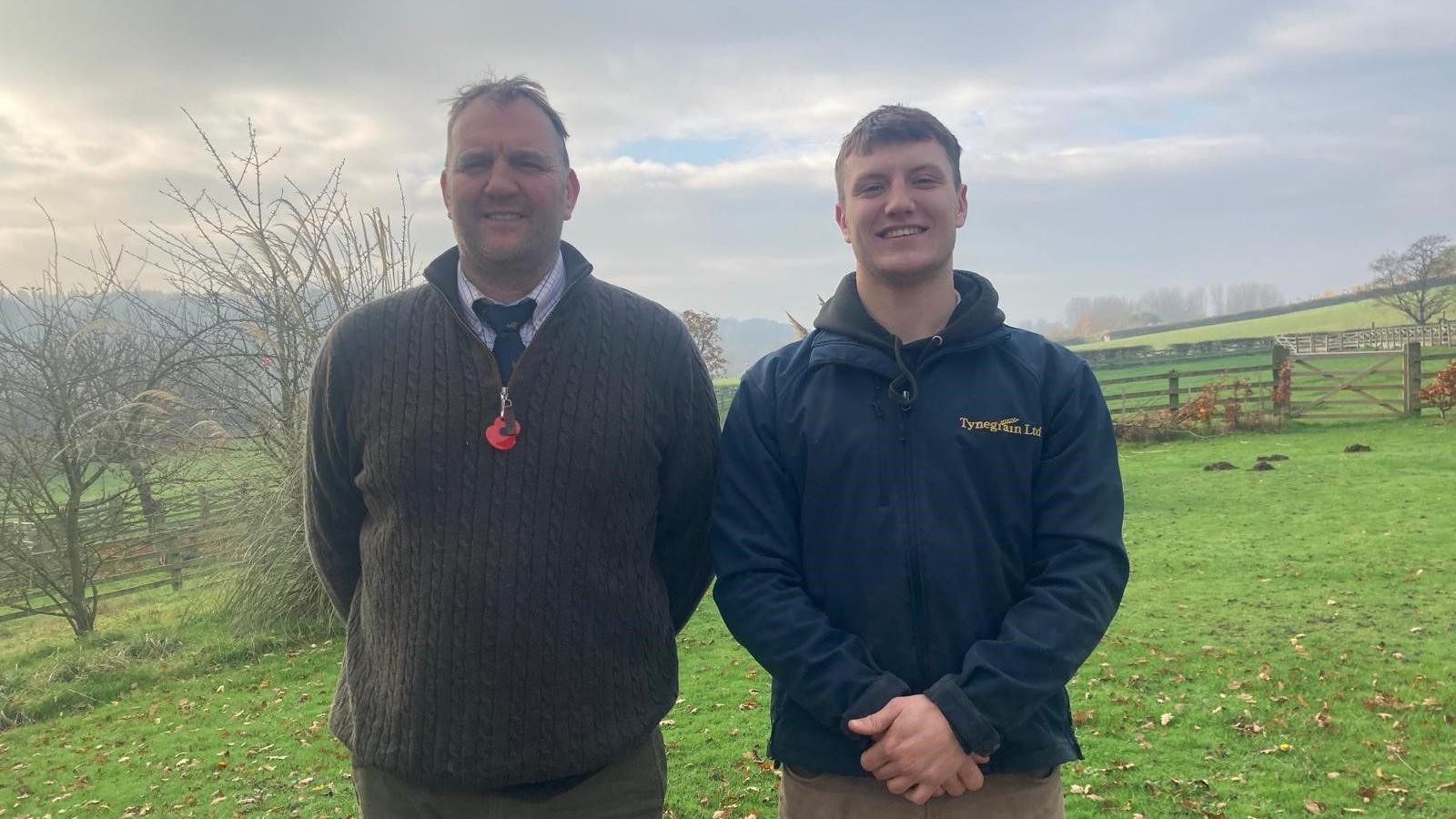 Andrew Moralee, who is in his 50s, stands next to his son Jack, who is 22. They are in a field with trees and a wooden fence behind. They are both smiling.  