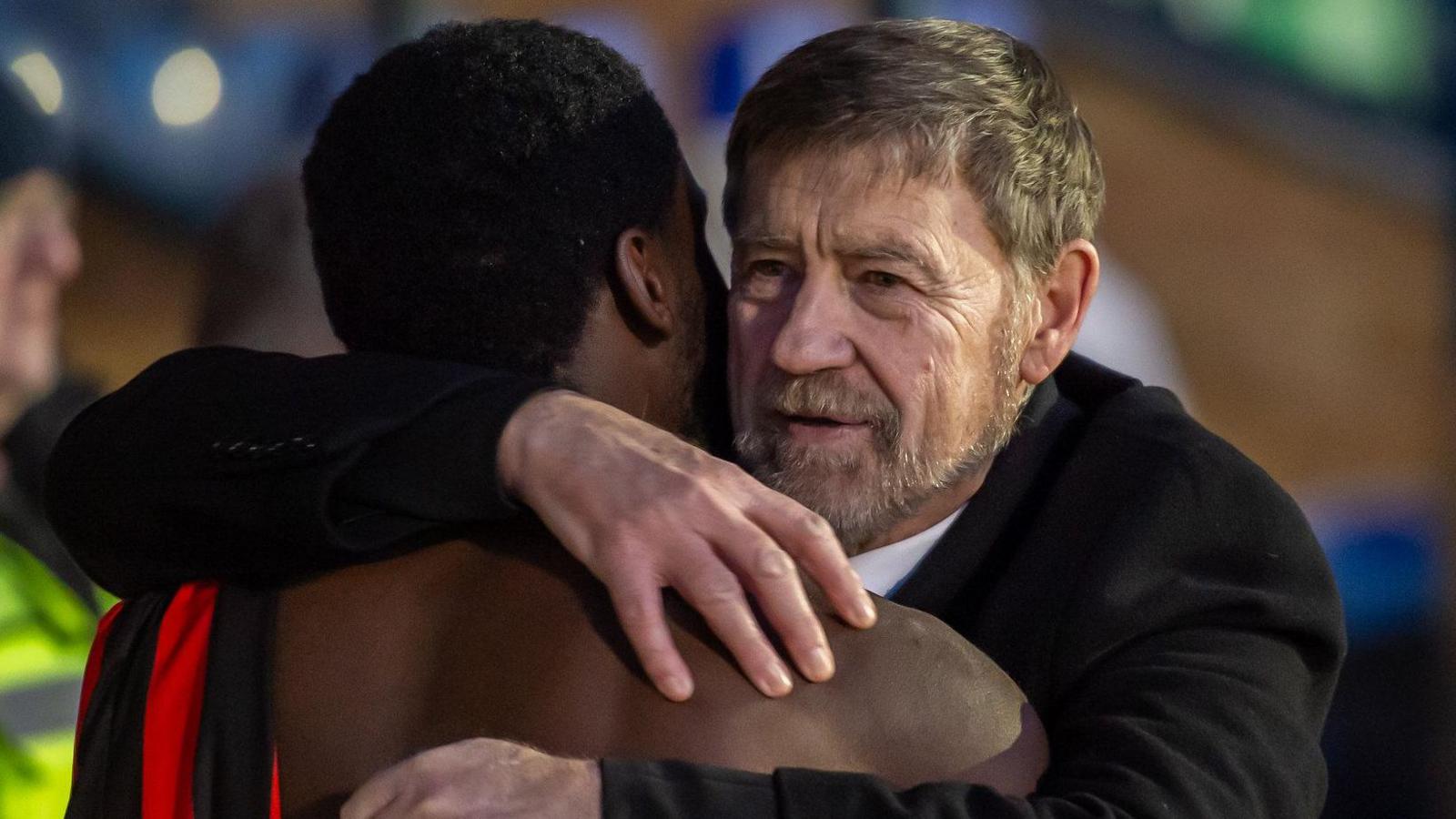 Maurice Dunk, with grey hair and a beard, hugs one of the players.