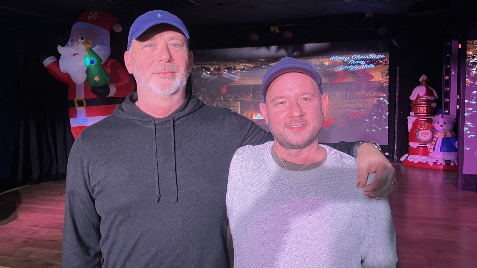 Close up image of Paul and Jason. The couple are standing next to each other looking into the camera. Paul has an arm around Jason. Paul is wearing a dark coloured hoodie and a blue cap. Jason is wearing a grey jumper with a blue cap. They are stood in the holiday park venue with large Christmas ornaments behind them.