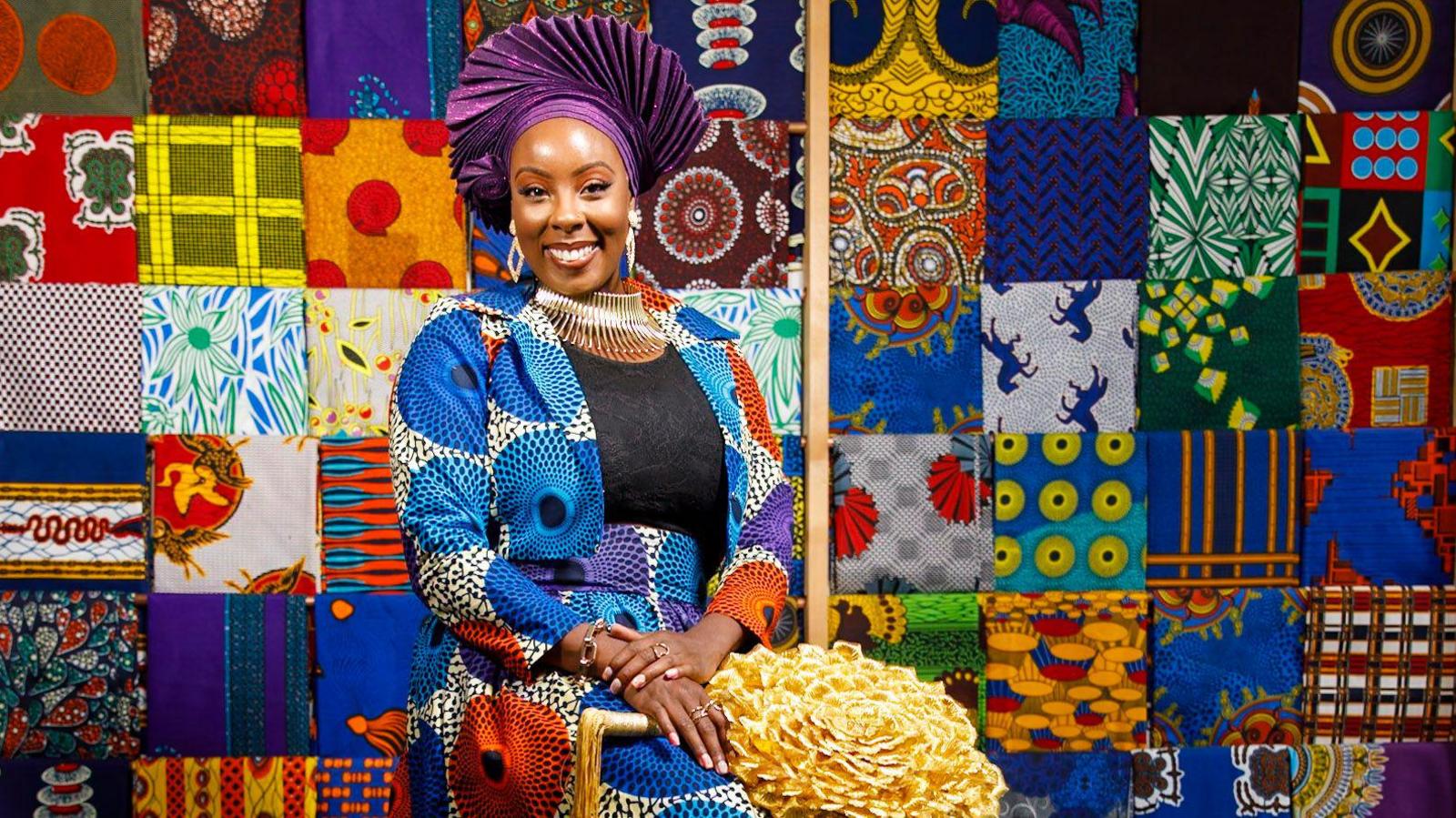 A woman in a bright purple fanned headdress and skirt and jacket with blue, purple and orange circular print, over a black top. She has an ornate silver collar necklace and dangly earrings. She is sitting down in front of a multi-coloured quilted display.