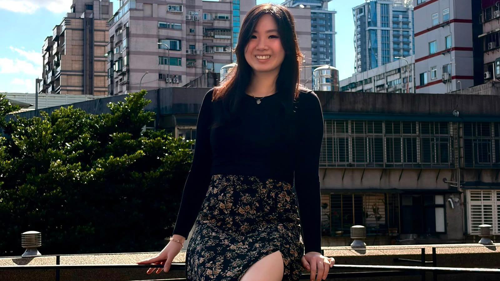 Young investor Michelle Huynh posing for a picture while sitting on a fence in front of a blue sky and a housing estate.