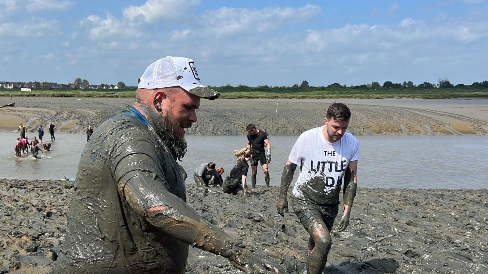 A muddy man wearing a peaked cap and behind him mud-splattered competitors climbing through the mud, Maldon